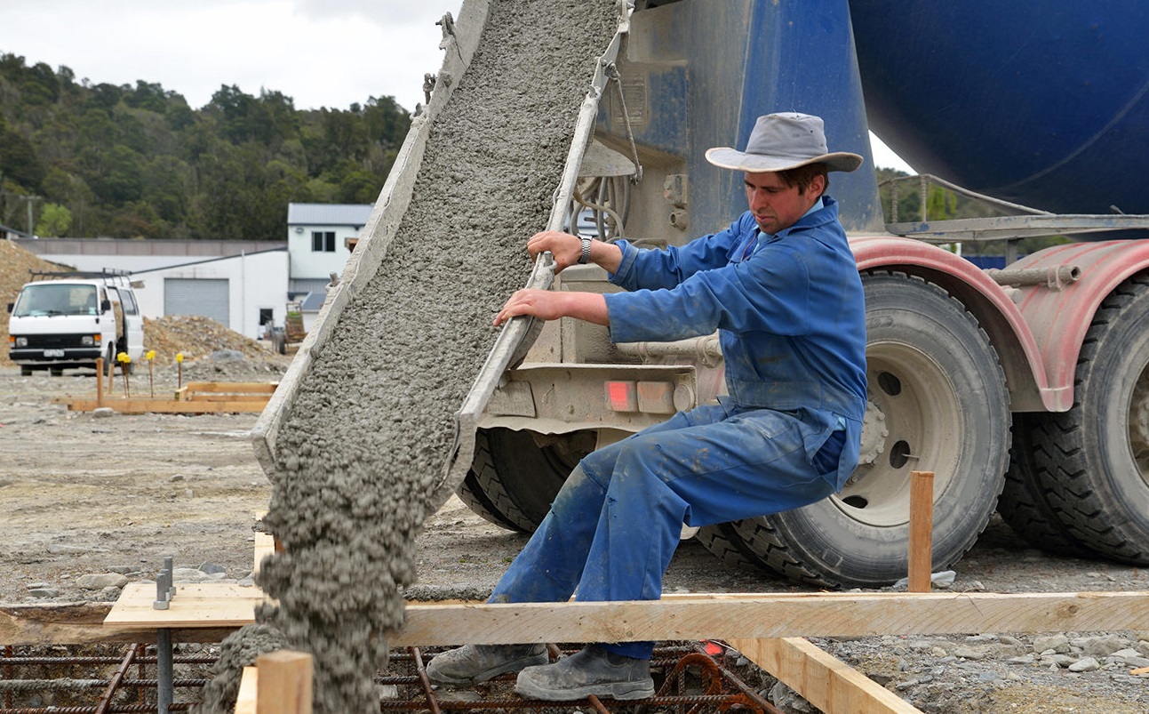 A builder directs wet concrete from a cement truck into the foundations of a large building.